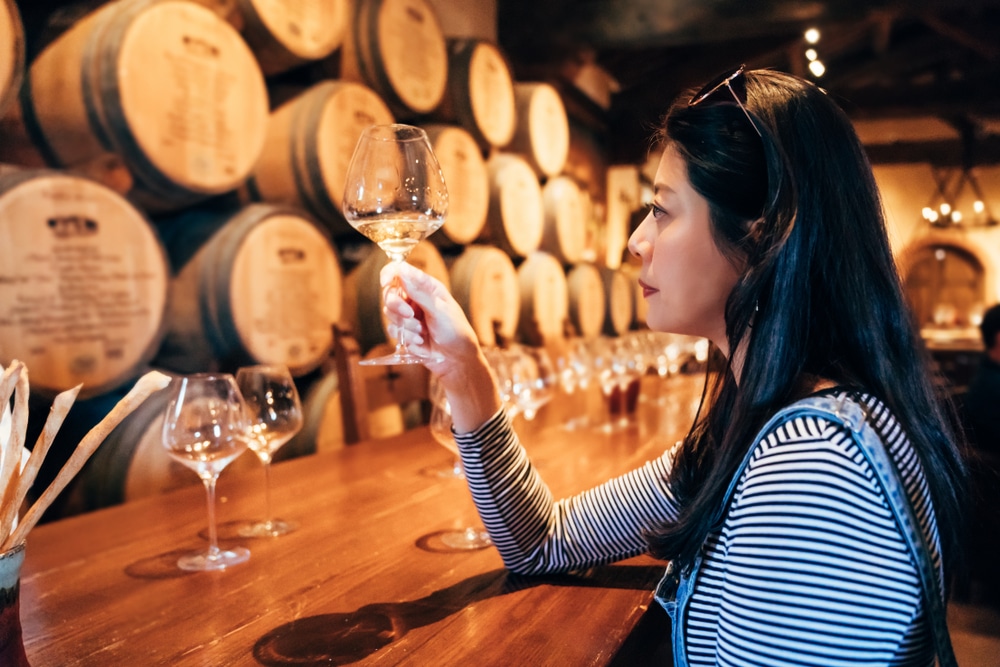 A woman examines a glass of wine while sitting at a wooden bar surrounded by wine barrels.
